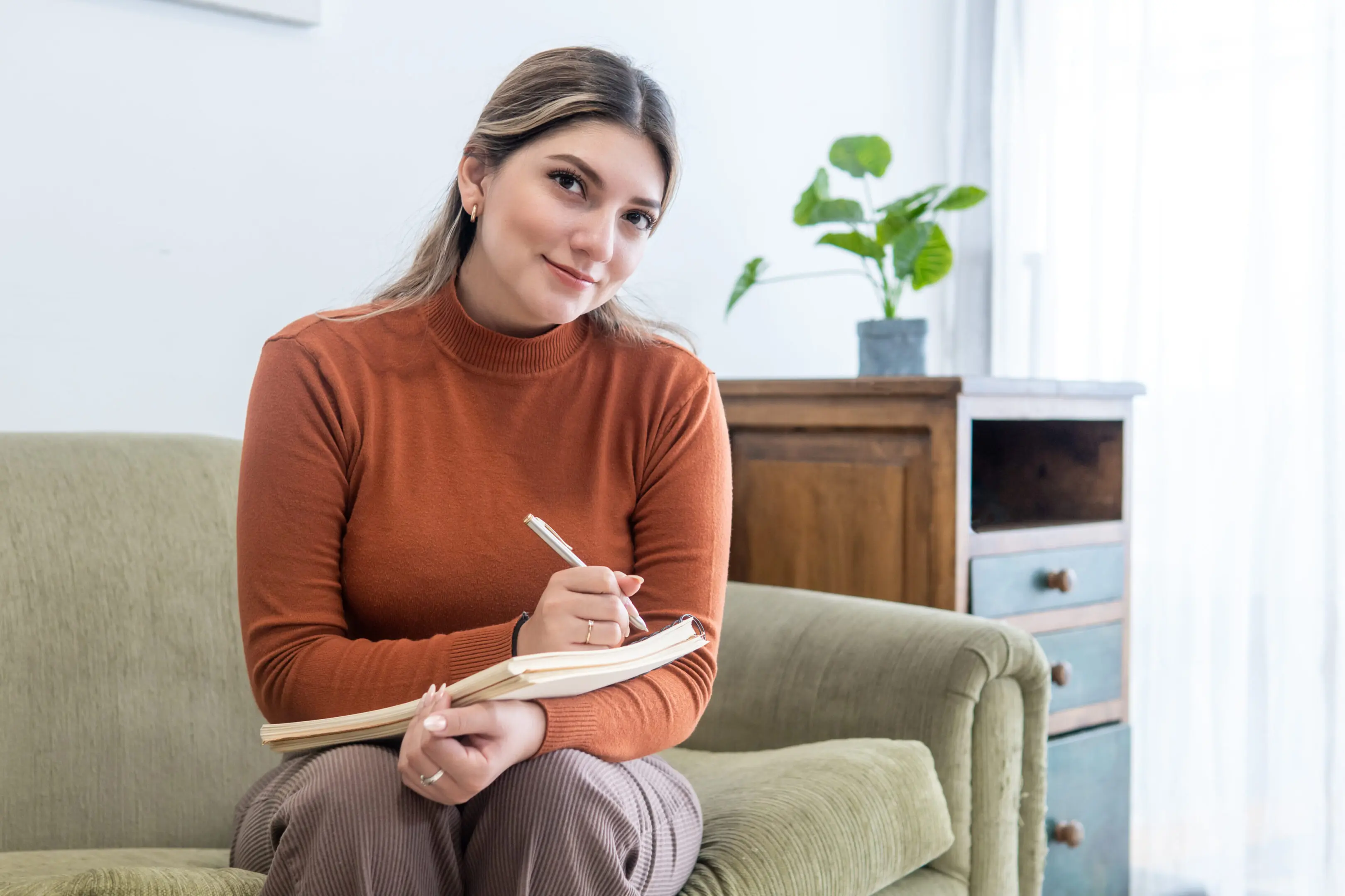 Woman sitting on a couch writing in a notebook with a pen.